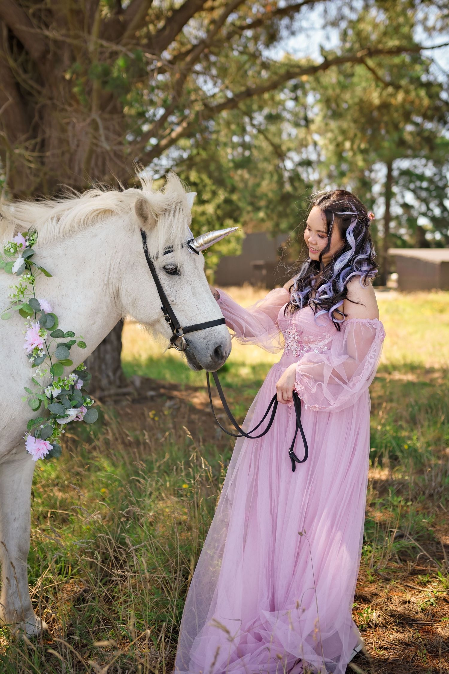 Woman in pink gown with a white unicorn decorated with flowers in a sunlit forest setting.