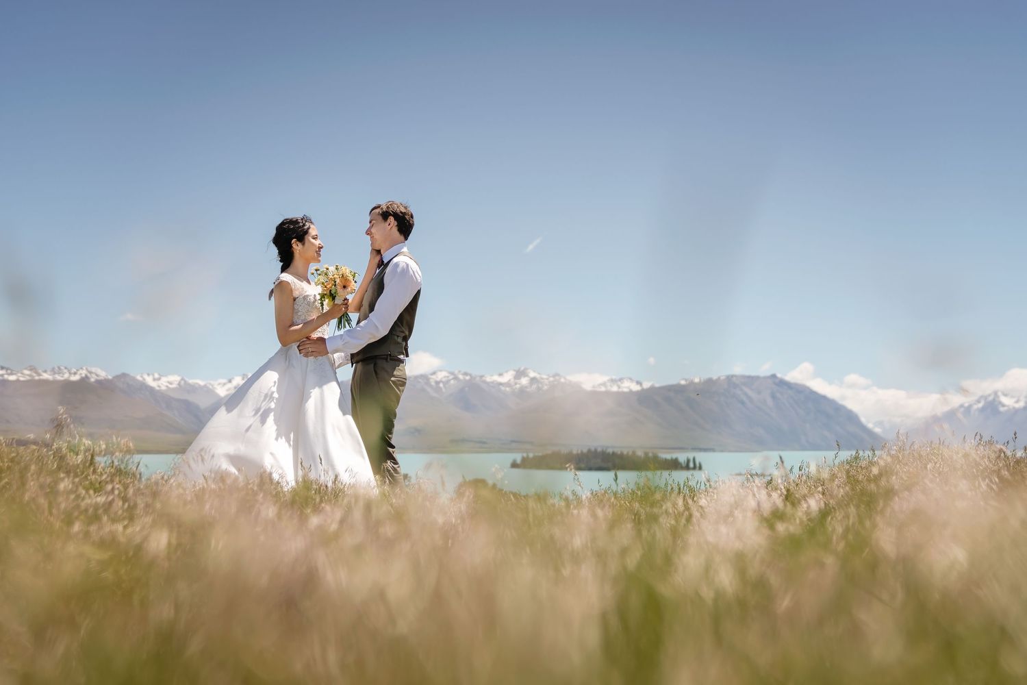 A couple in wedding attire stands together in a scenic mountain meadow with tall grass and peaks in the background.