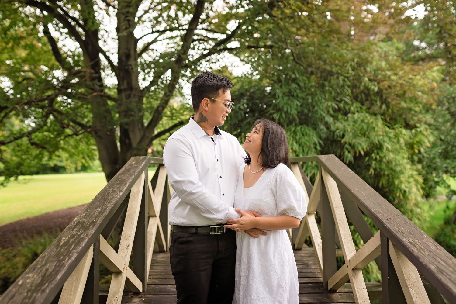 Couple sharing a romantic moment on a wooden bridge surrounded by lush green trees in a park.