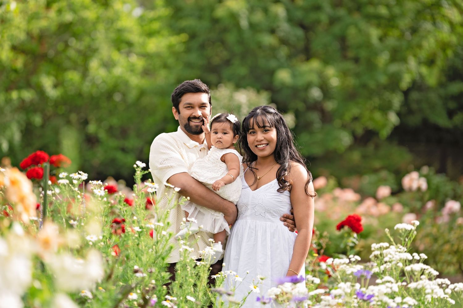 Family of three posing among colorful blooming flowers in a lush garden, parents holding their young child.