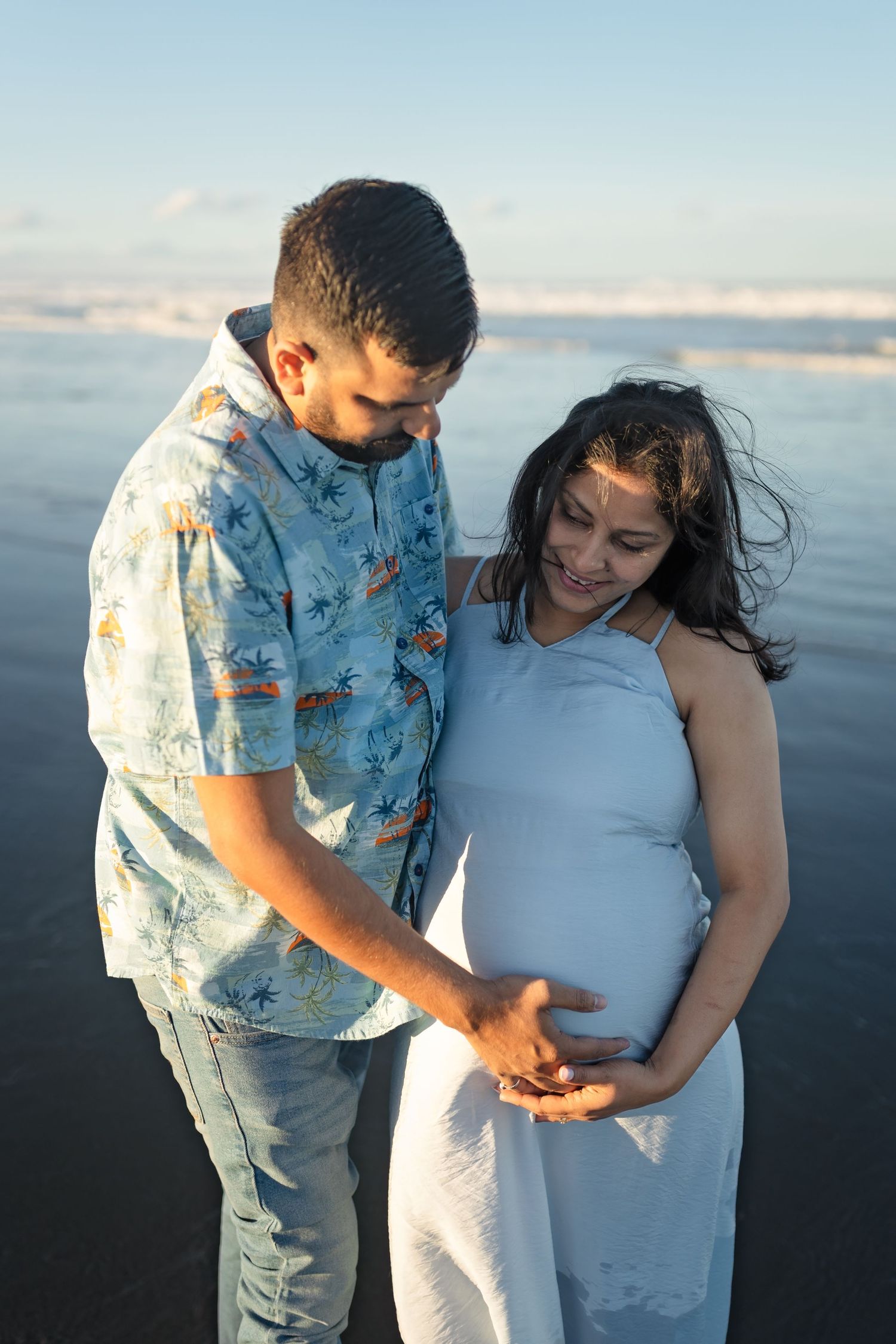 A couple in white attire shares emotional moments during a beach maternity photoshoot at sunset.