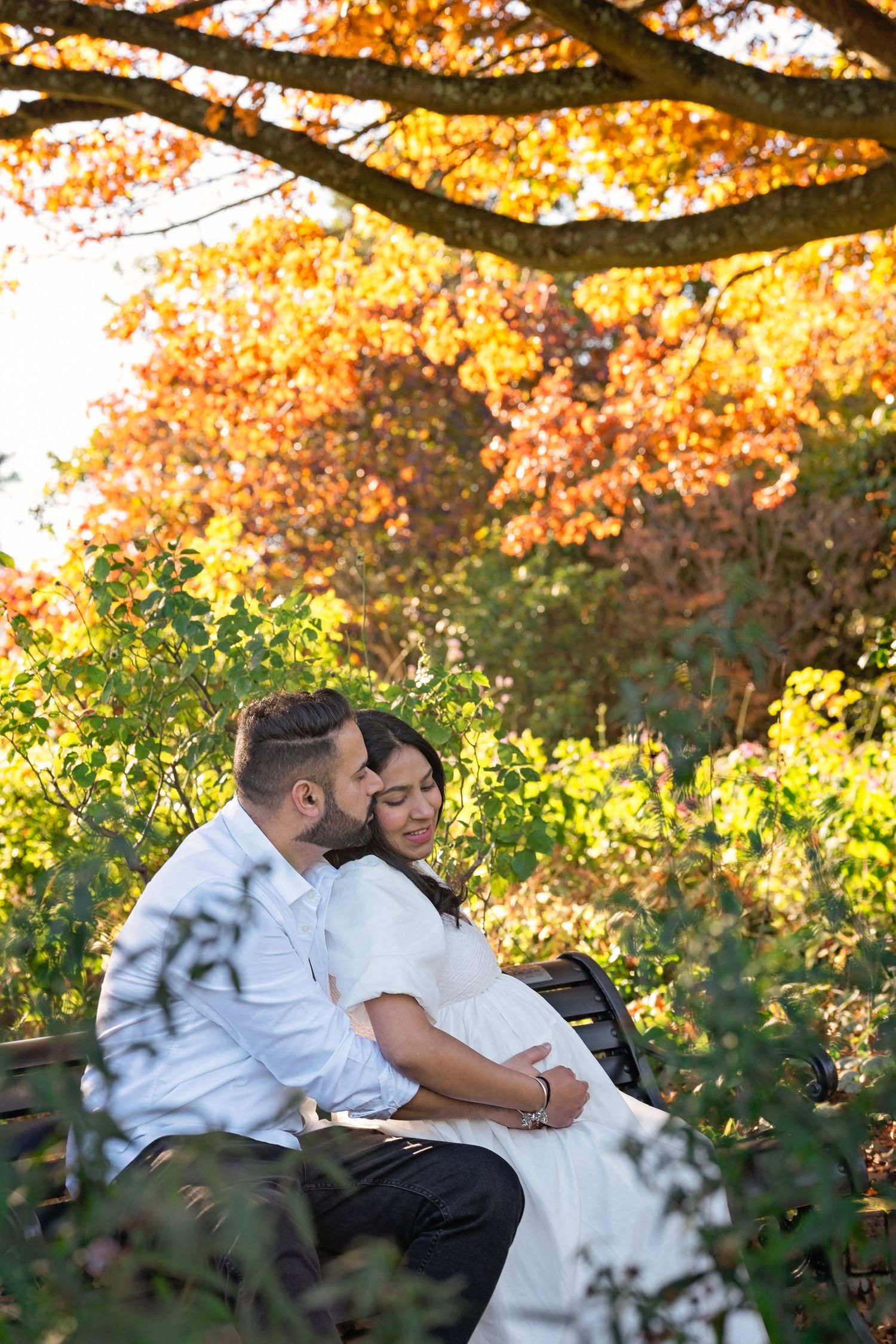 A couple shares a tender moment on a bench surrounded by golden autumn foliage in a garden setting.