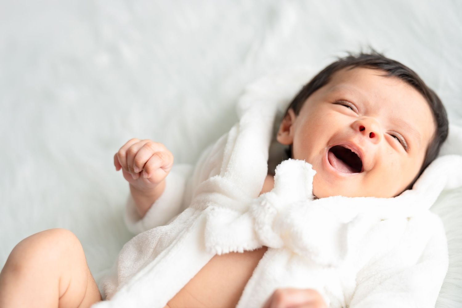 Laughing baby in white outfit lying on white background during photo shoot.