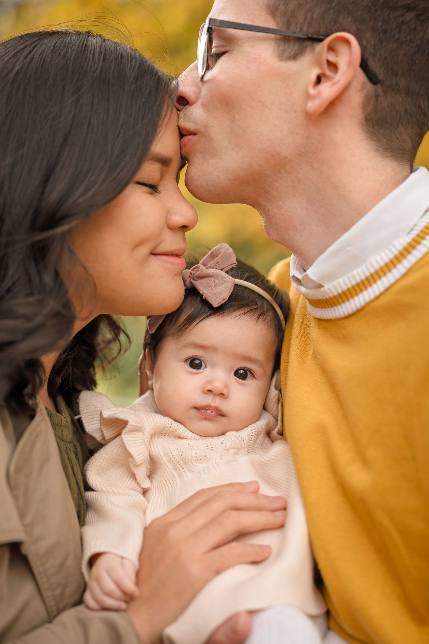 A couple kisses while holding their infant daughter in a warm autumn outdoor setting.