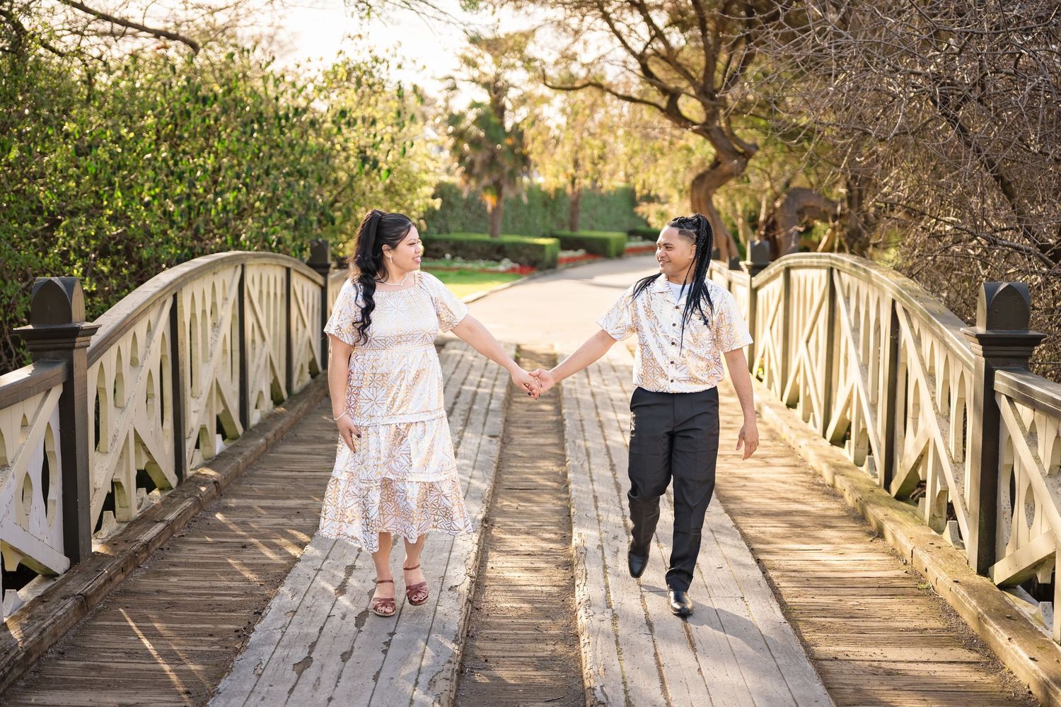 Couple holding hands on a wooden bridge in a lush garden, woman in white lace dress, man in black pants.