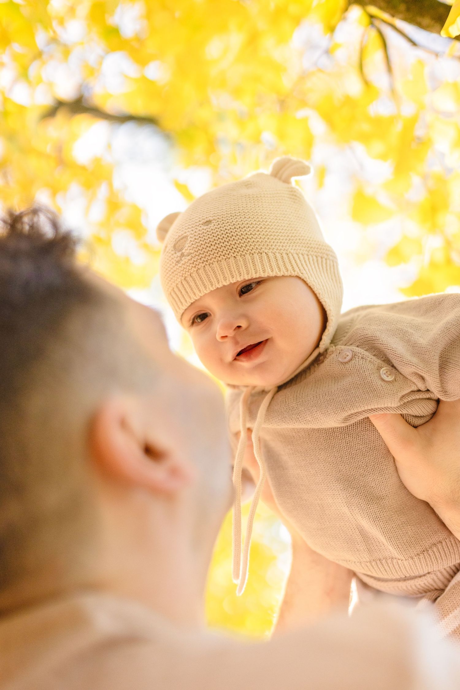 Baby wearing a beige knit hat smiles under golden autumn foliage in soft natural lighting.