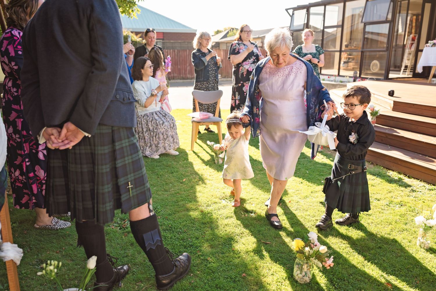 Wedding guests gathering on green lawn with elderly woman walking with young children during outdoor ceremony.