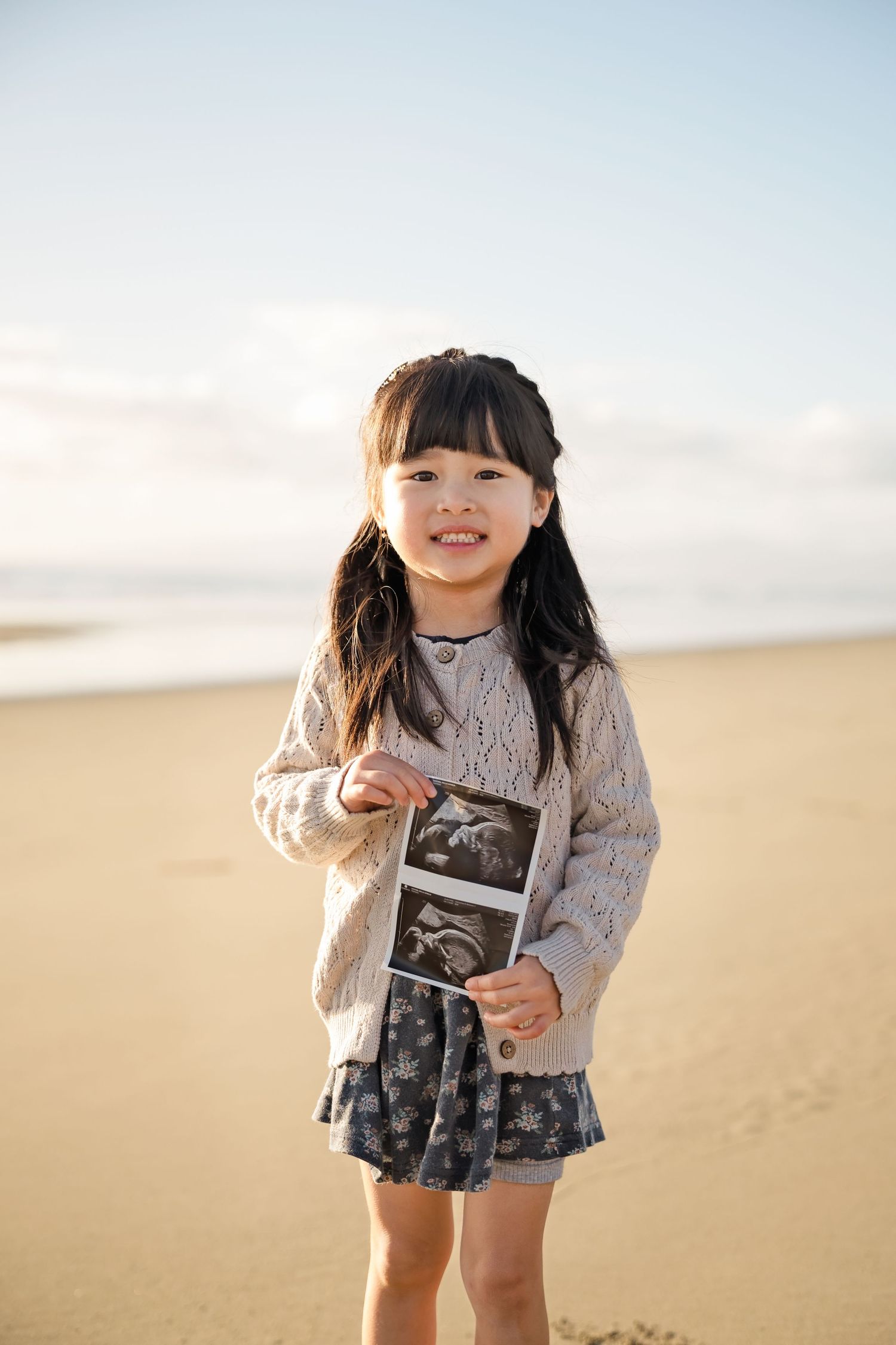 Young Asian girl smiling on sandy beach holding ultrasound photo, wearing floral skirt and knit cardigan.