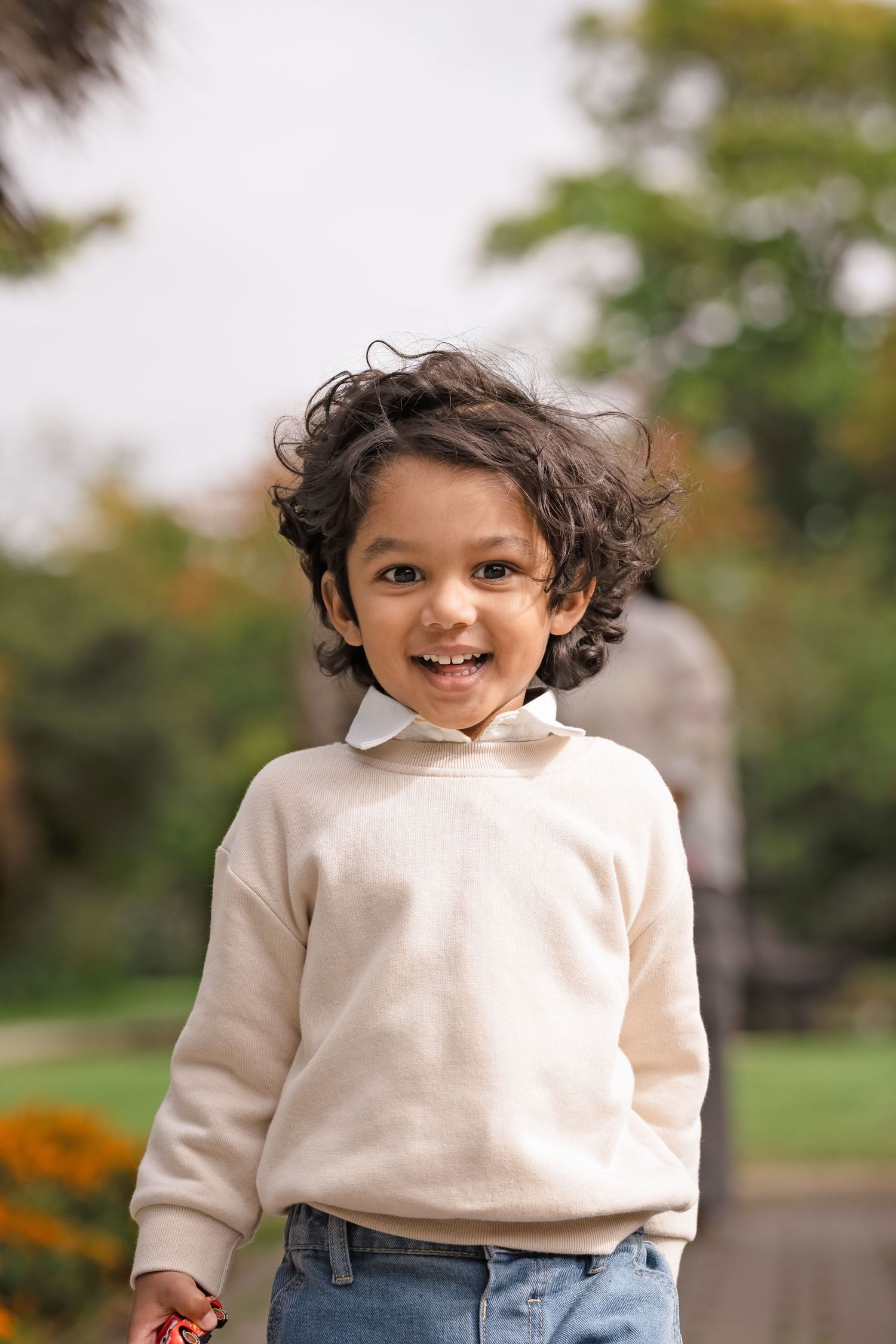 A happy toddler with curly hair wearing a beige sweater and jeans smiles outdoors in a park setting.