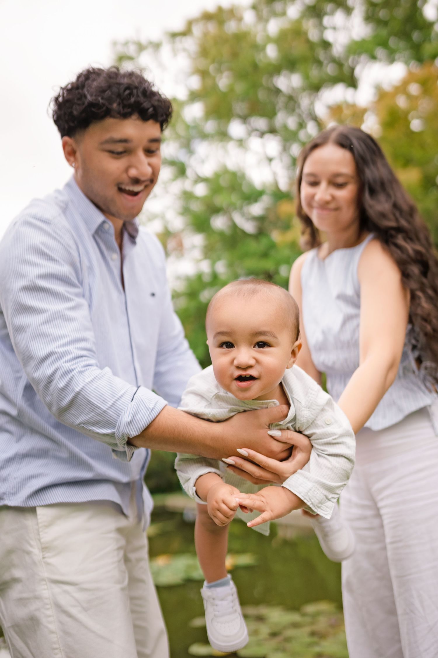 Parents playfully lifting smiling baby outdoors near pond, family portrait in natural setting.