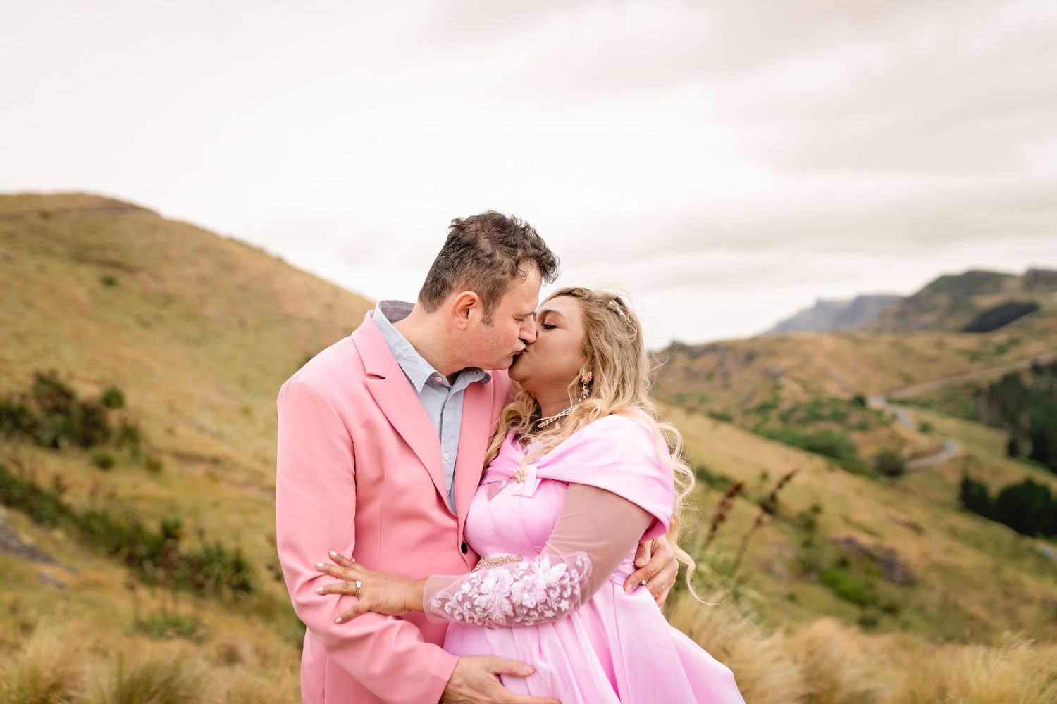 Couple in matching pink outfits sharing a romantic kiss on a scenic mountain hillside under a cloudy sky.