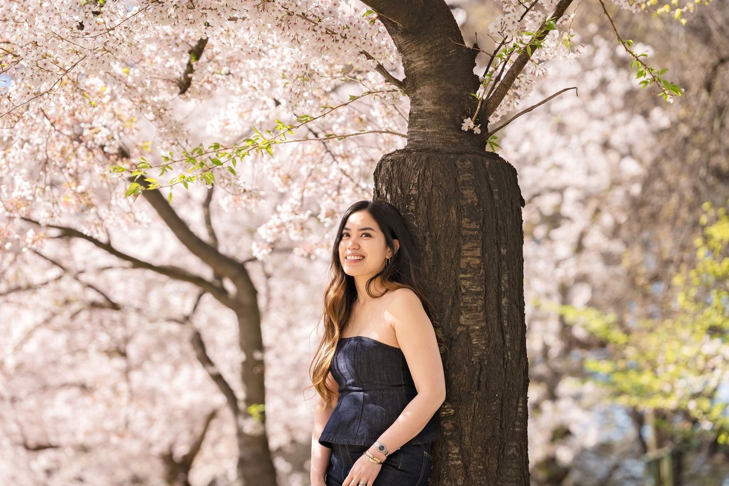 Woman in black strapless dress leaning against tree surrounded by blooming cherry blossoms in spring.