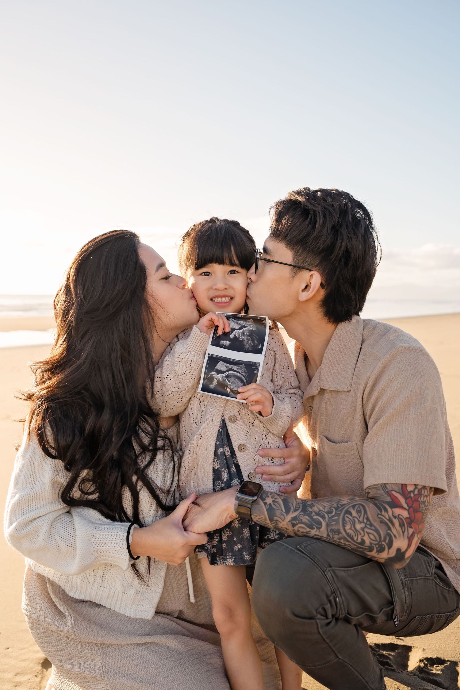 Happy couple kissing their smiling toddler holding an ultrasound photo on a sunny beach.