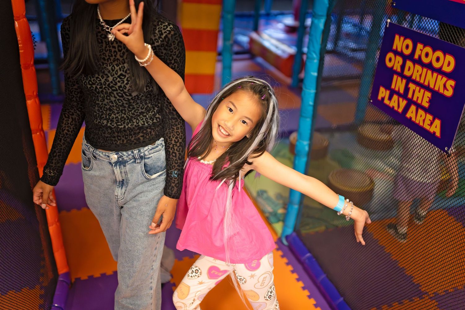 A young girl in a pink top poses playfully in an indoor play area with a no food or drinks sign visible.