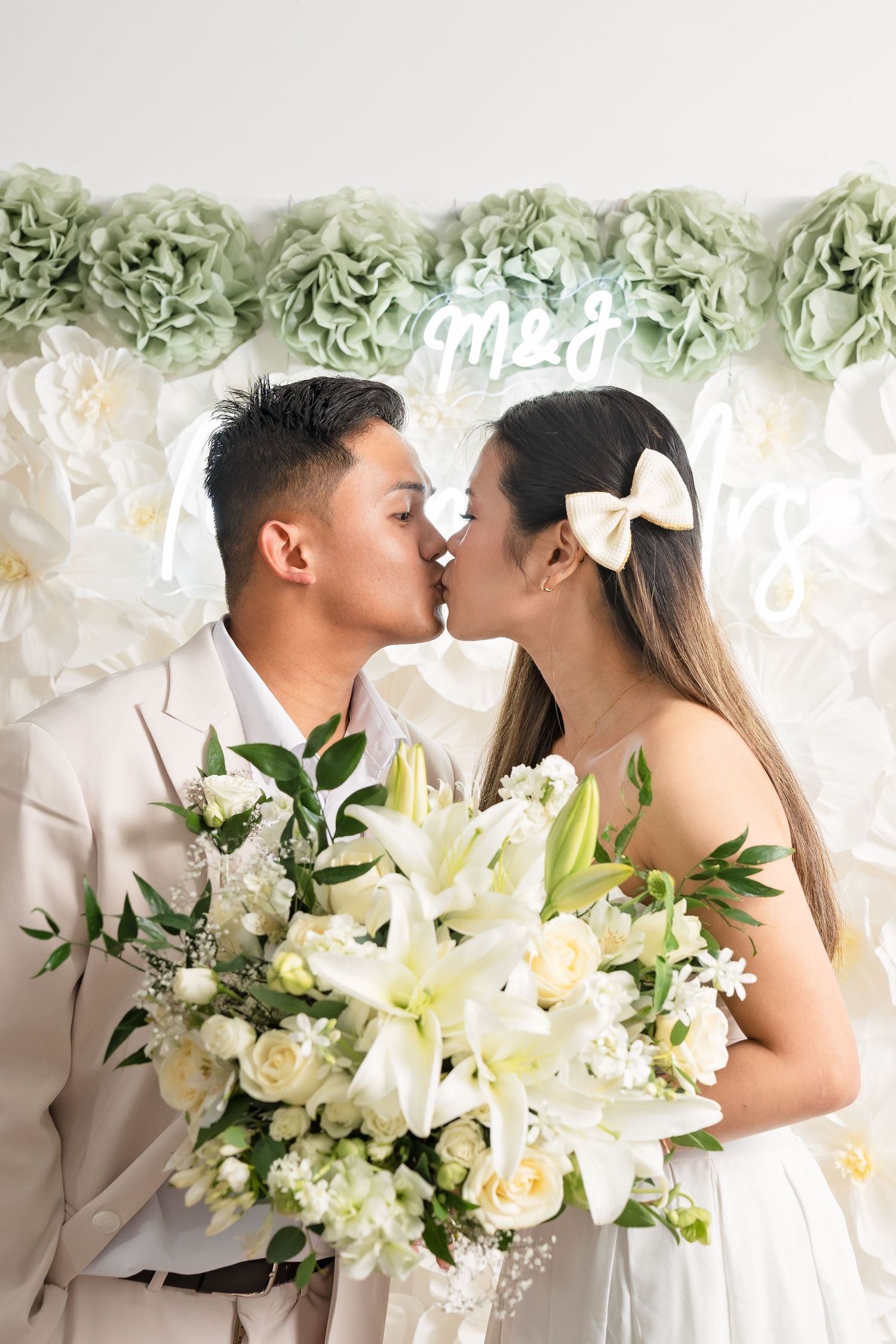 Newlywed couple shares a kiss holding white lily bouquet in front of decorative floral backdrop.