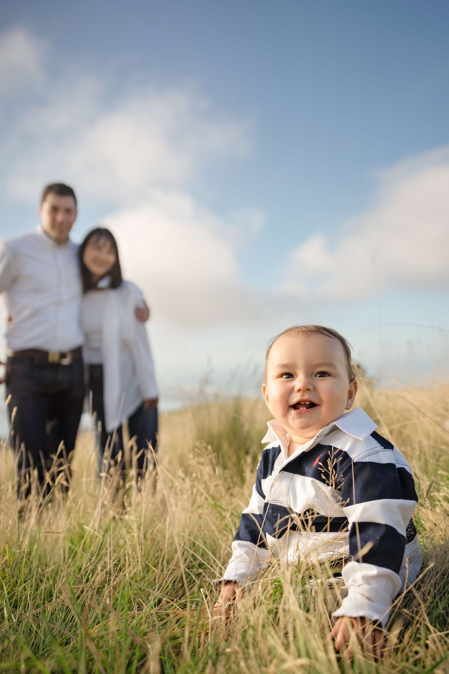 Happy baby in striped outfit sits in tall grass while parents stand together under blue sky in a warm family outdoor portrait.