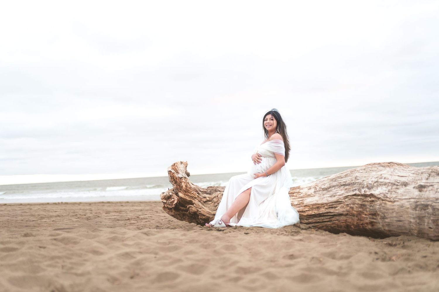Maternity portrait of woman in white dress seated on driftwood at a coastal beach.