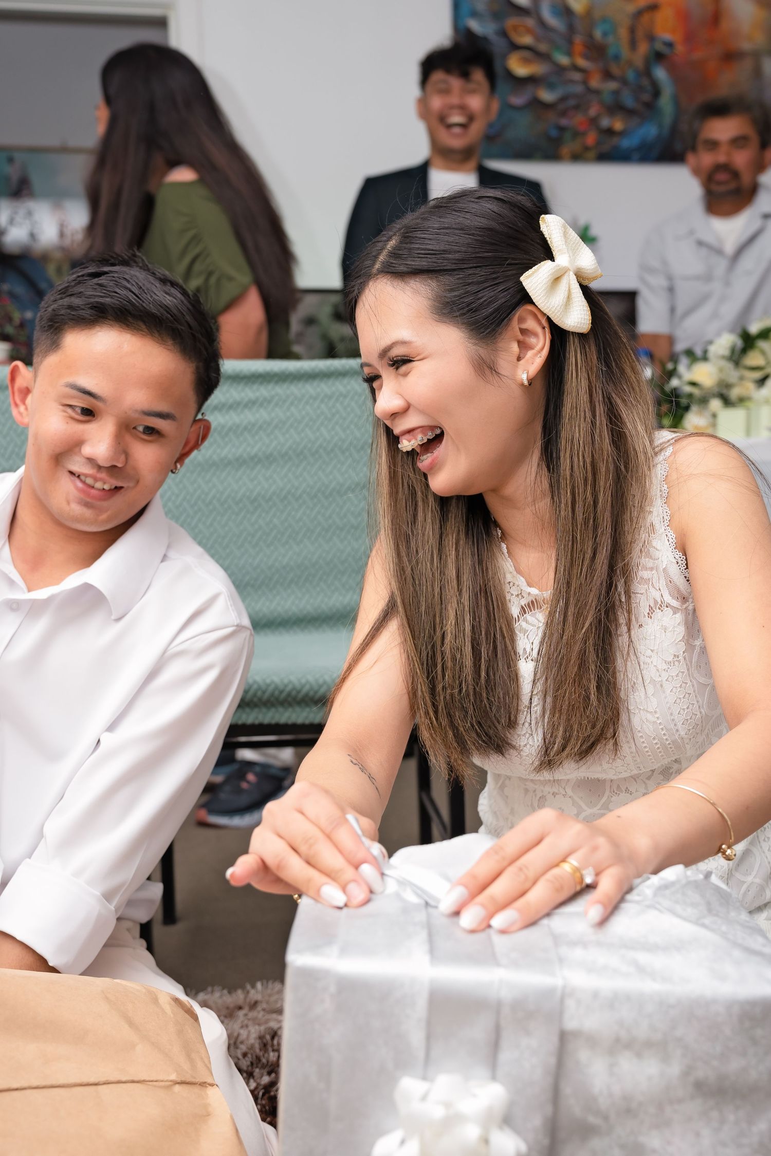 A joyful couple opens wedding gifts together, smiling and laughing at a celebration with guests in the background.