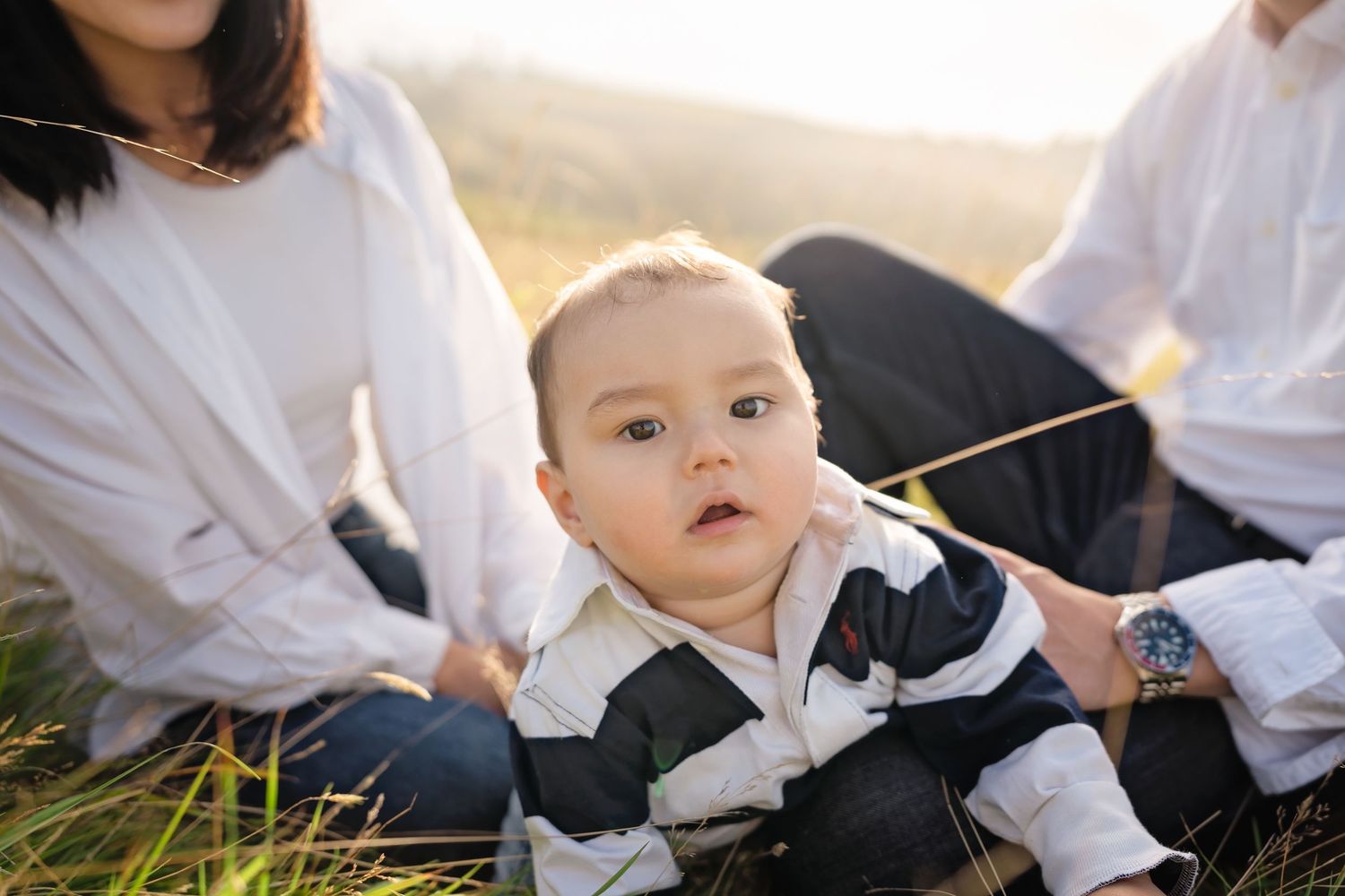 Adorable baby in striped outfit sitting in grass with family during golden hour outdoor photoshoot.