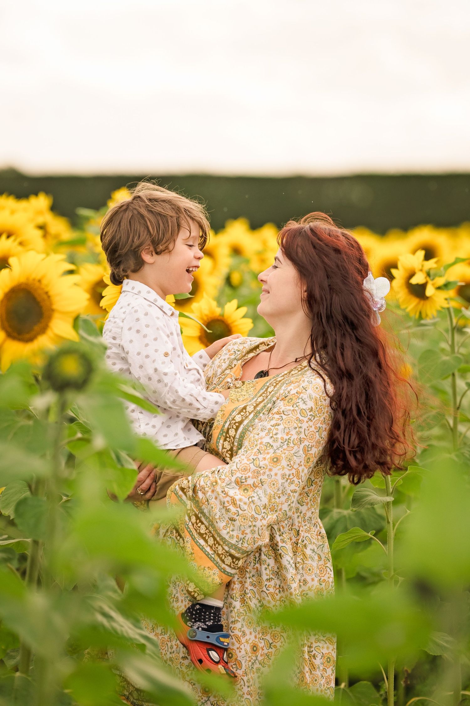A joyful mother lifts her laughing son in a vibrant sunflower field, both wearing floral outfits on a cloudy day.