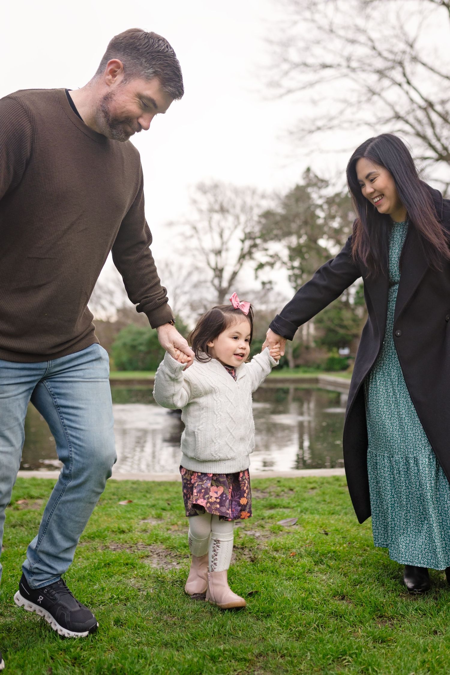 A family plays together on grass near a pond, holding hands and enjoying a sunny day in the park.