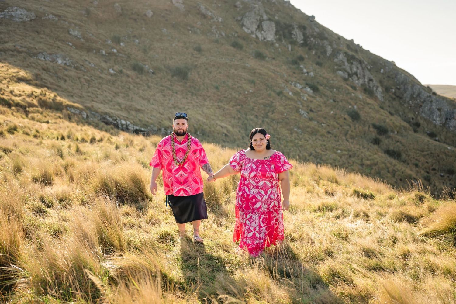 Couple in matching pink outfits walking hand in hand on a grassy hillside with rocky mountains behind them.