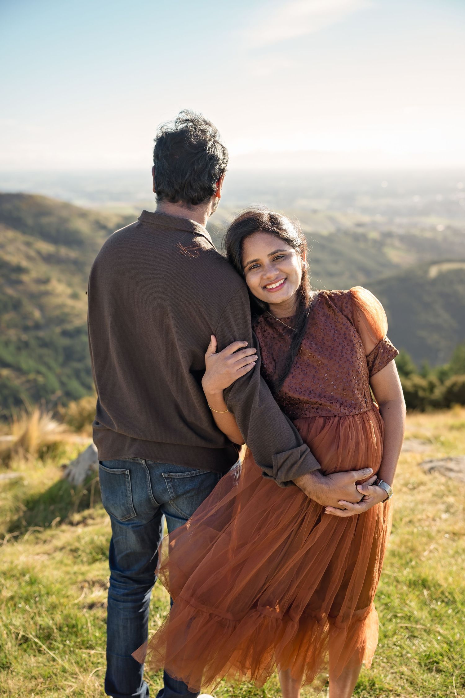 Smiling woman in brown dress embracing man on scenic hilltop with mountain valley views.