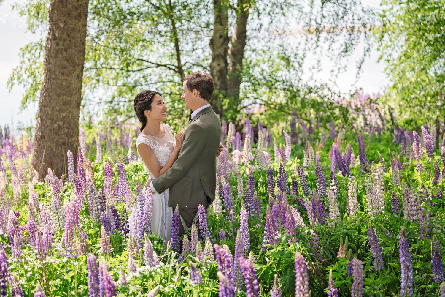 Bride and groom embracing in a vibrant field of purple and pink lupine flowers surrounded by trees.