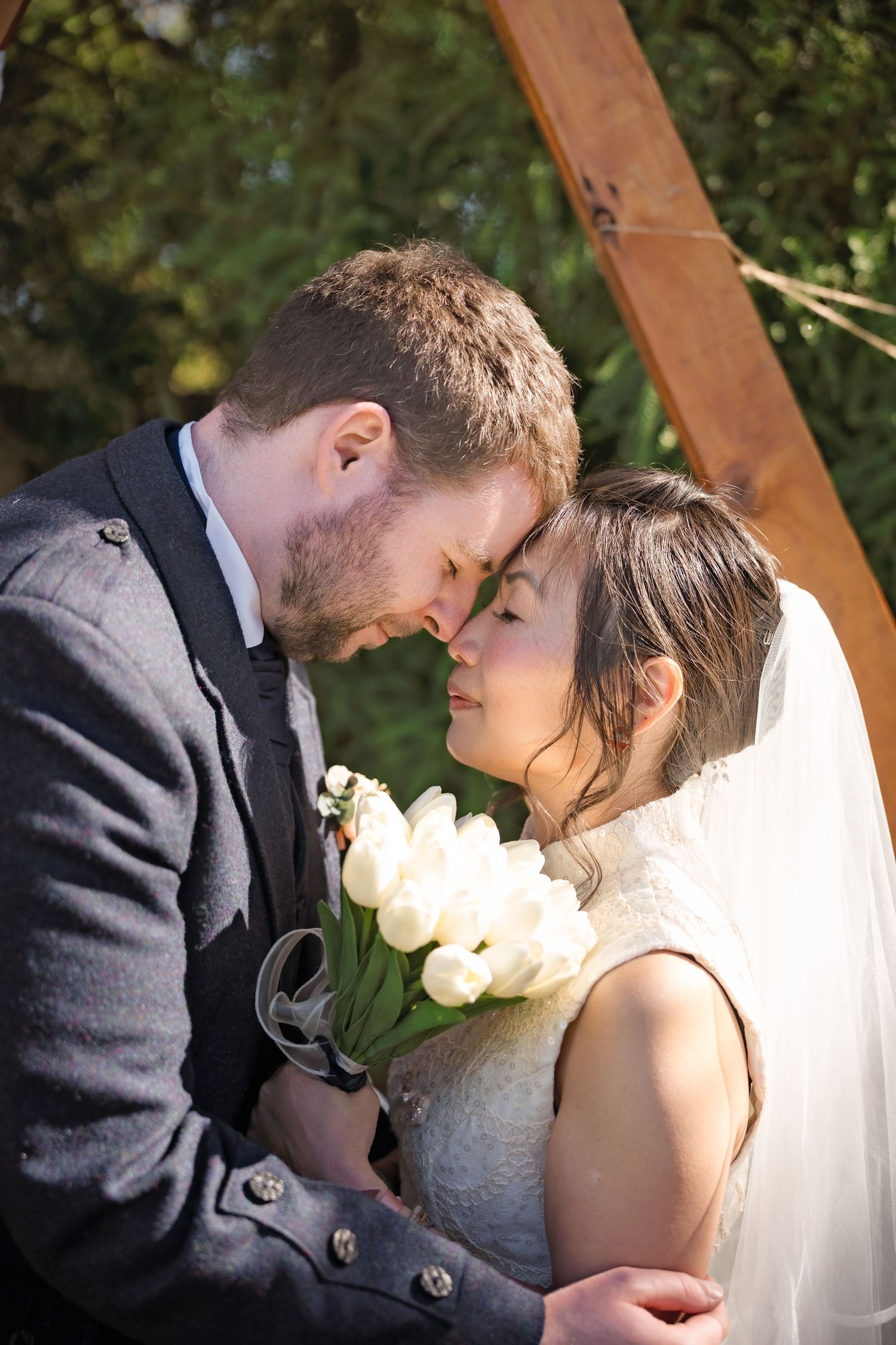 Bride and groom touch foreheads romantically under wooden arch, bride holding white tulip bouquet.
