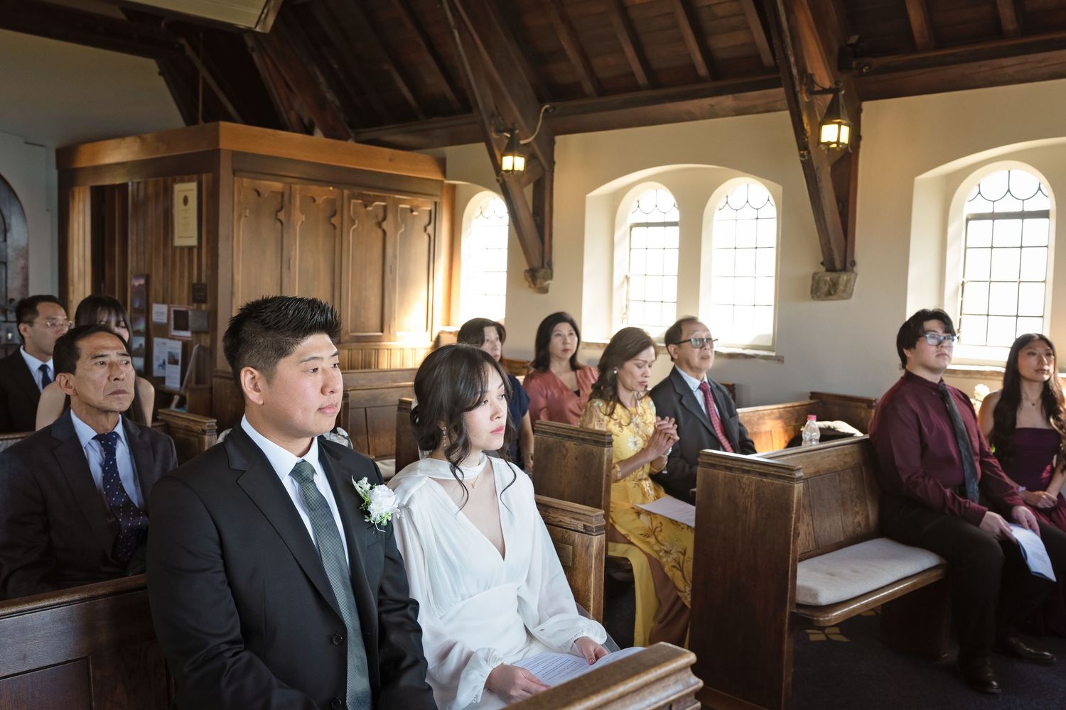 Asian wedding couple and guests seated in a charming wooden chapel during an intimate wedding ceremony.