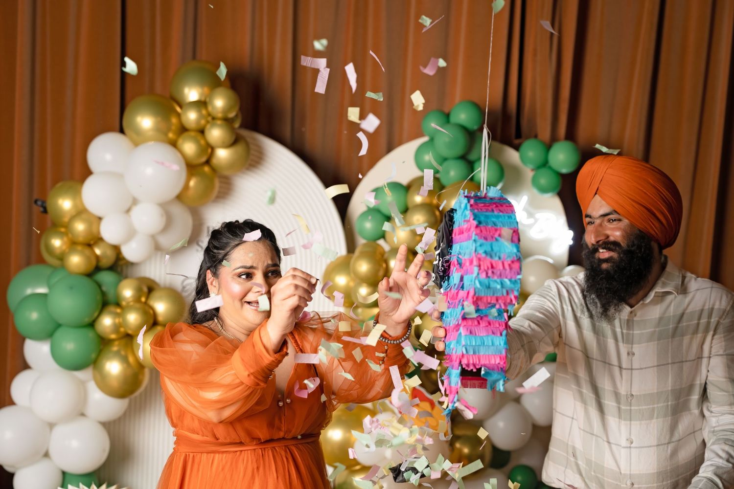 Sikh couple celebrating with confetti and a pinata at a baby shower with green and gold balloon decorations.
