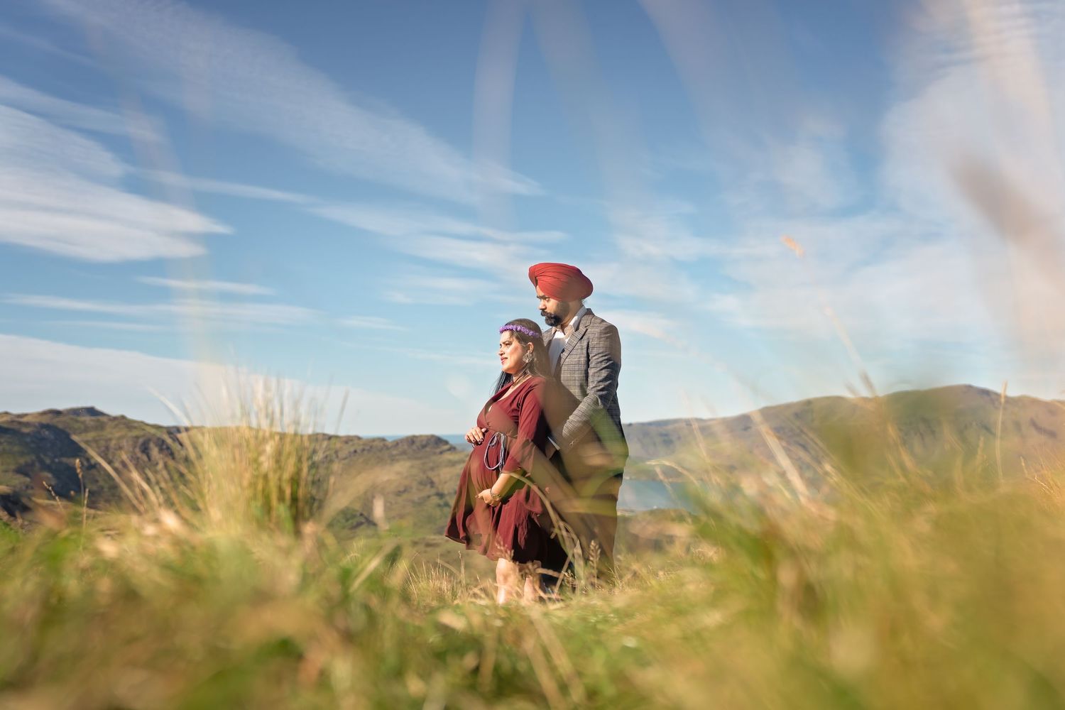 A couple in warm clothing embraces while standing on a scenic hilltop with mountains and blue sky in the background.