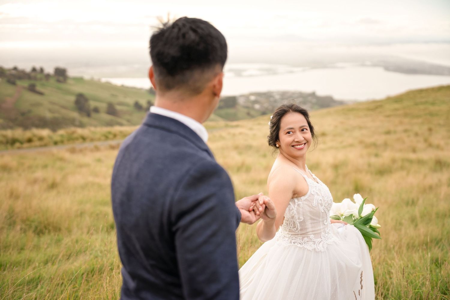 Bride in lace gown holds groom's hand on green hillside, smiling back with scenic coastal views behind them.