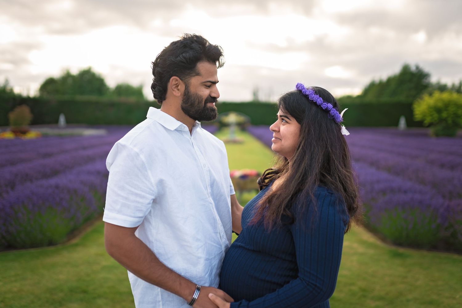 Expecting couple shares an intimate moment in a blooming lavender field under cloudy skies.