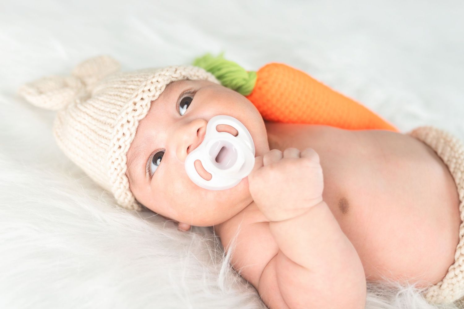 Baby wearing knit beanie hat and pacifier lies next to decorative carrot on soft white blanket.