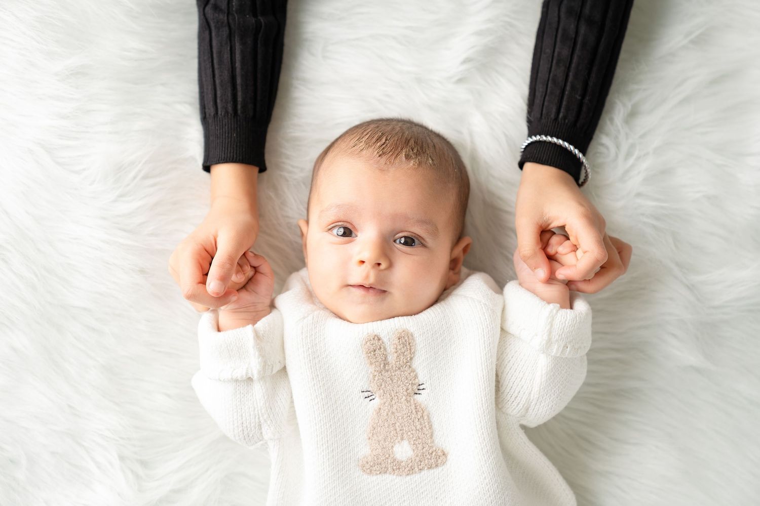 A baby wearing a white bunny sweater lies on white fabric while holding two adult hands.
