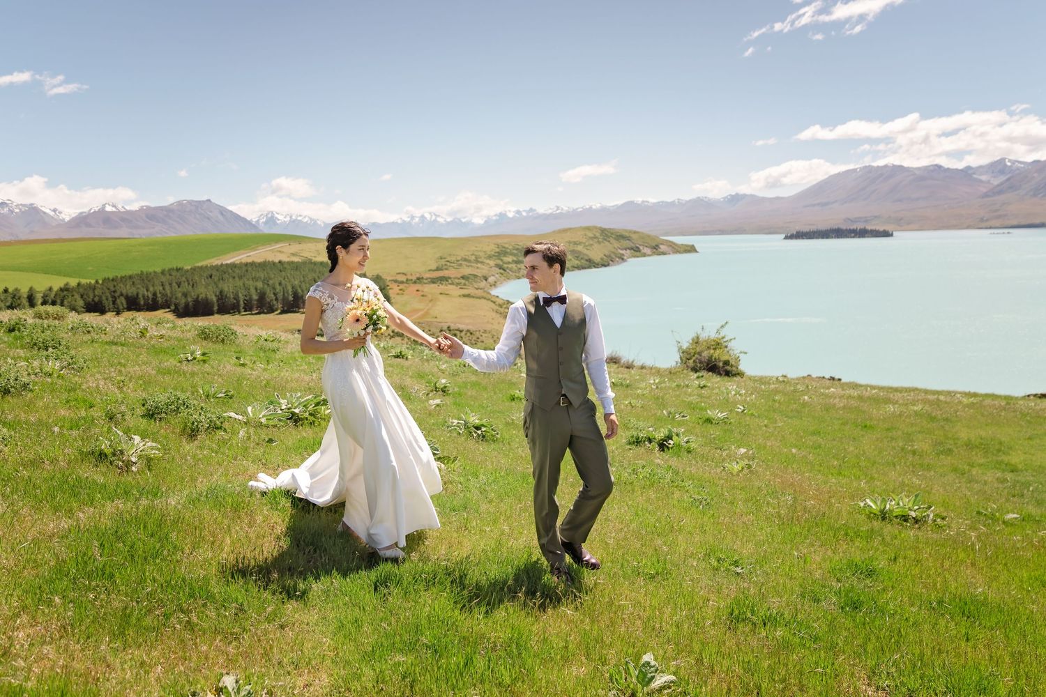 Bride and groom walking hand in hand on green hillside overlooking turquoise Lake Tekapo, New Zealand.