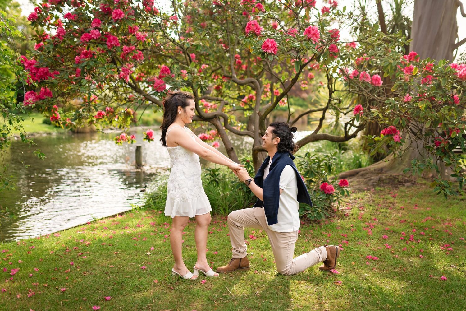 Couple sharing a romantic marriage proposal moment in a garden with blooming pink flowers by a serene pond.