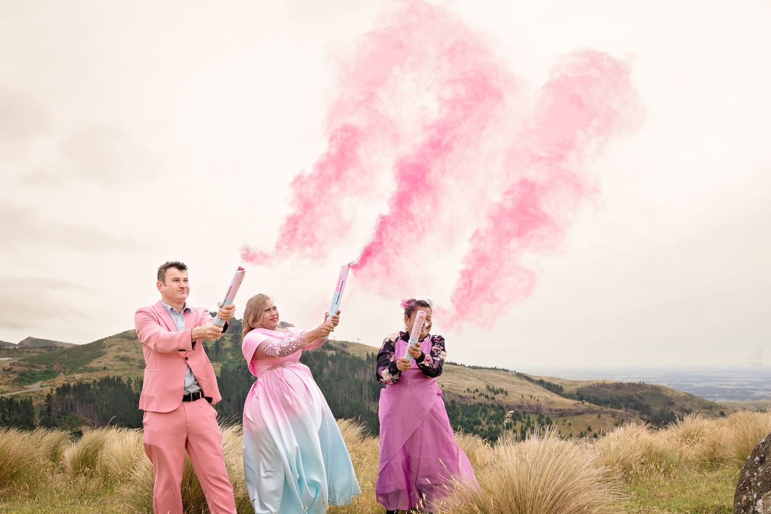 Three people celebrate a gender reveal with pink smoke bombs outdoors, wearing pink and pastel outfits against a mountain backdrop.