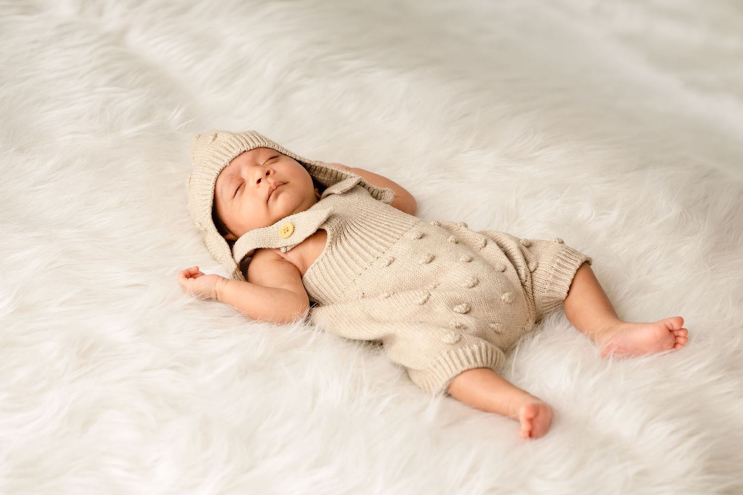 Sleeping newborn baby in beige knit romper and bonnet hat lying on white fluffy fur blanket.