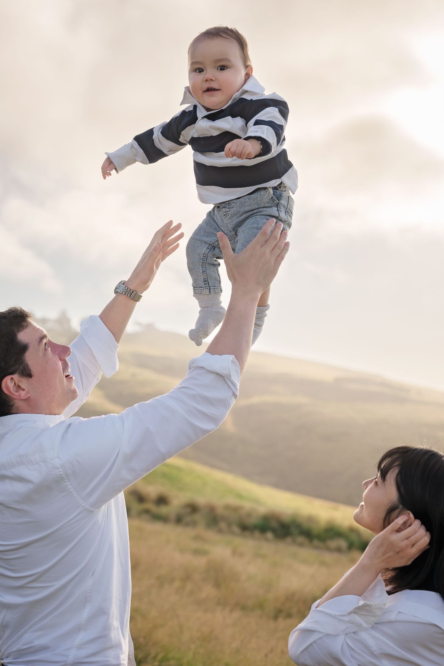 Joyful parents toss their baby in the air outdoors in a scenic grassy hillside setting.