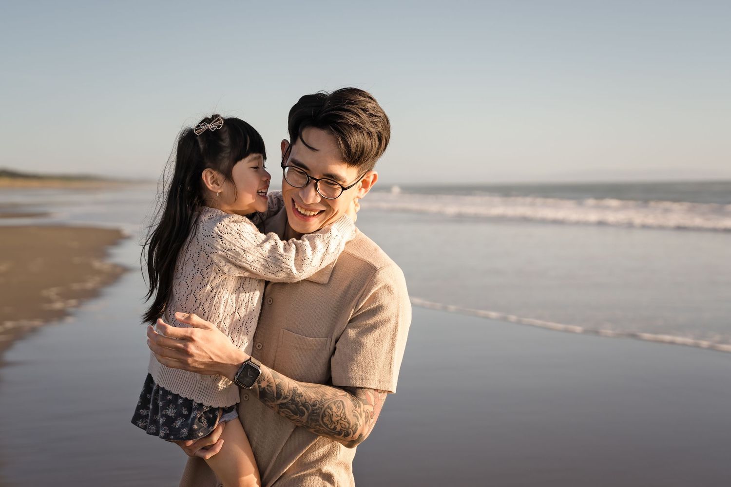 A smiling man with tattoos holds a young girl on a sunny beach, sharing a joyful embrace by the ocean.