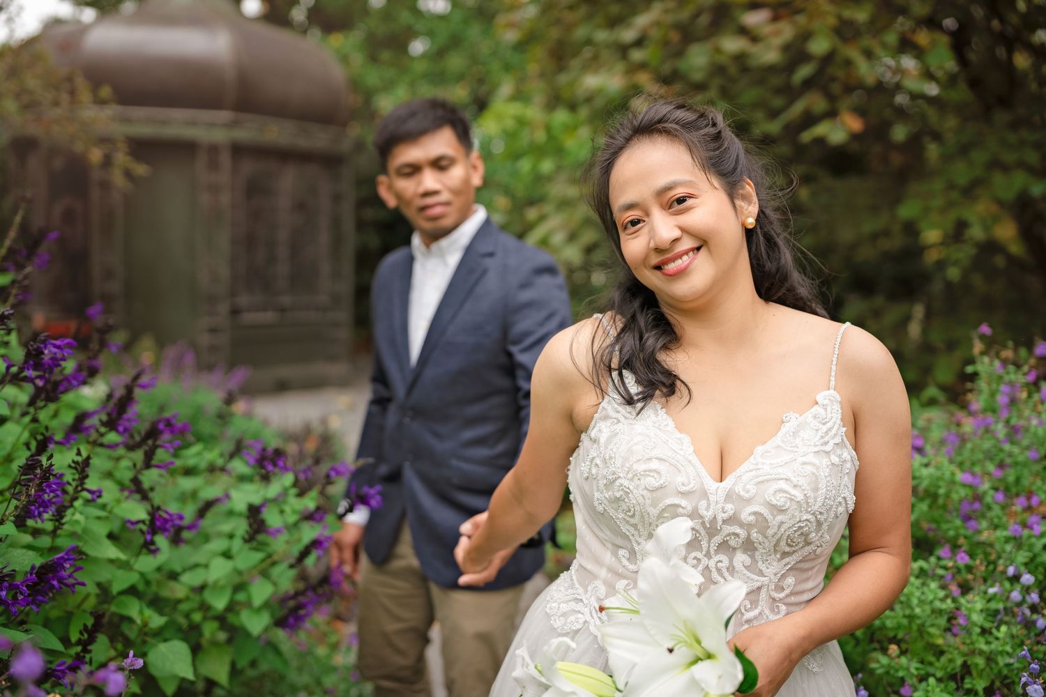 Bride in lace gown holds white lilies while groom stands behind her among purple flowers in a garden setting.
