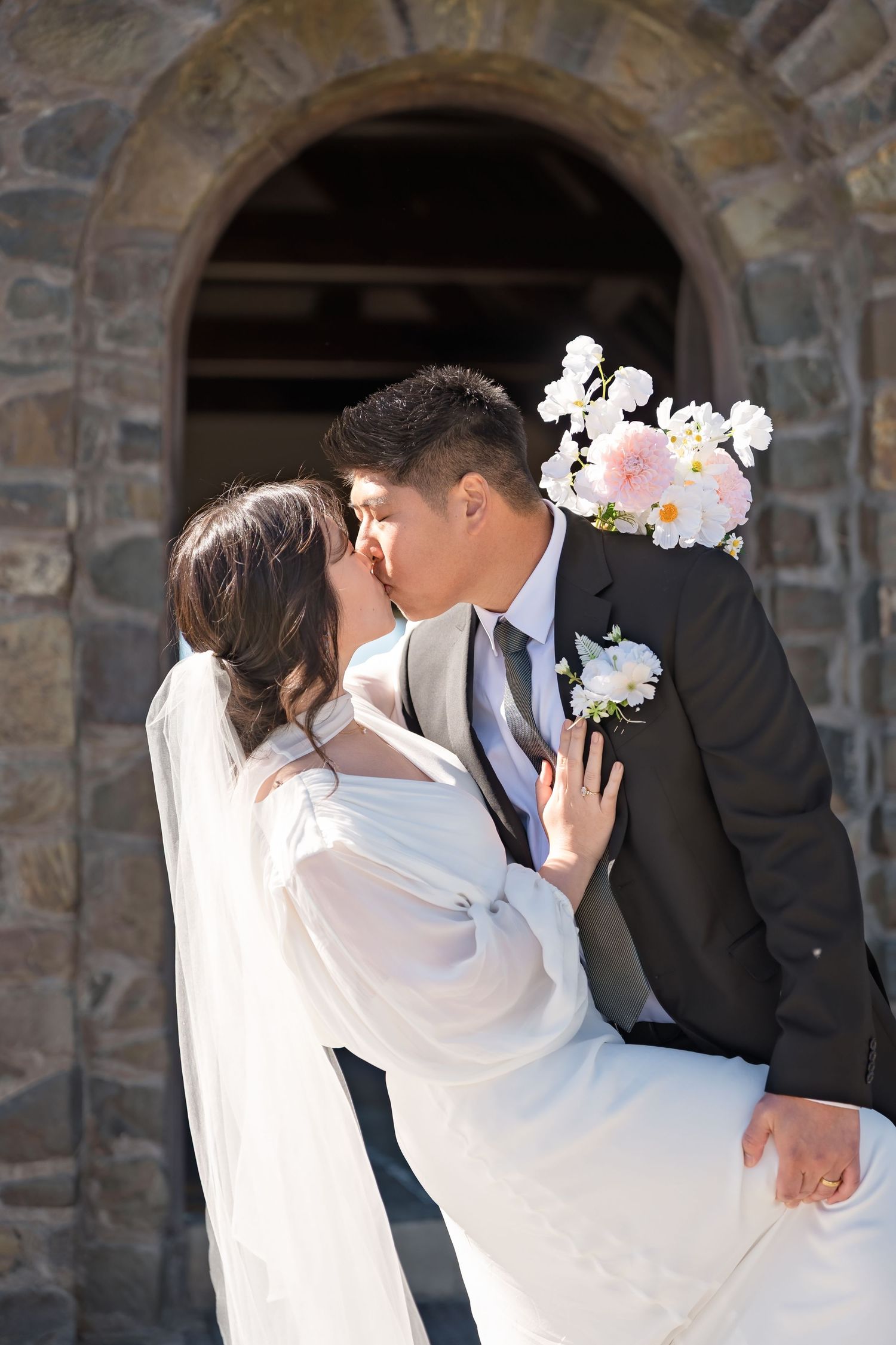 Newlywed couple shares a romantic kiss outside a rustic stone church on their wedding day.