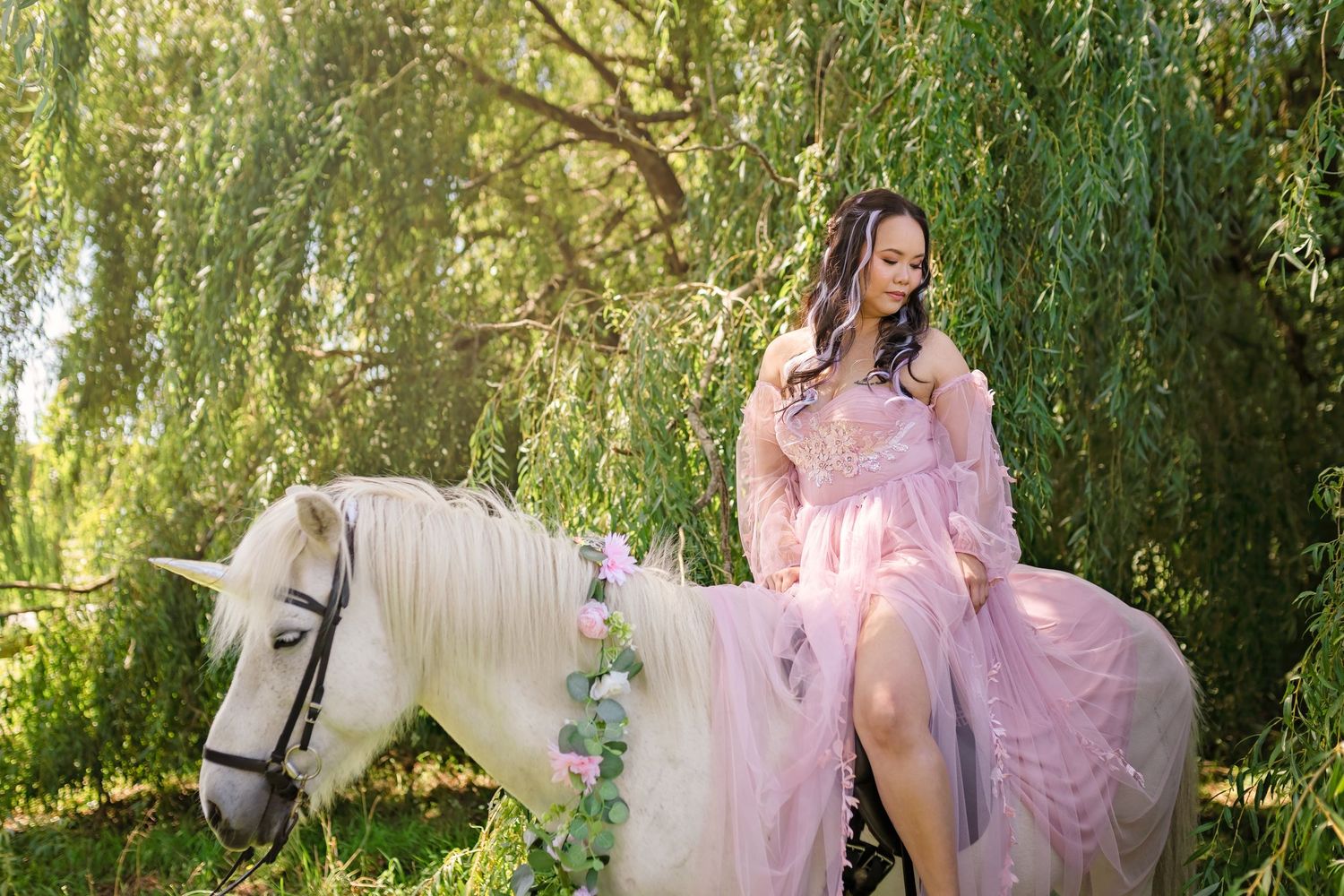 Woman in pink gown riding a white unicorn horse under weeping willow trees in a fairy tale fantasy setting.