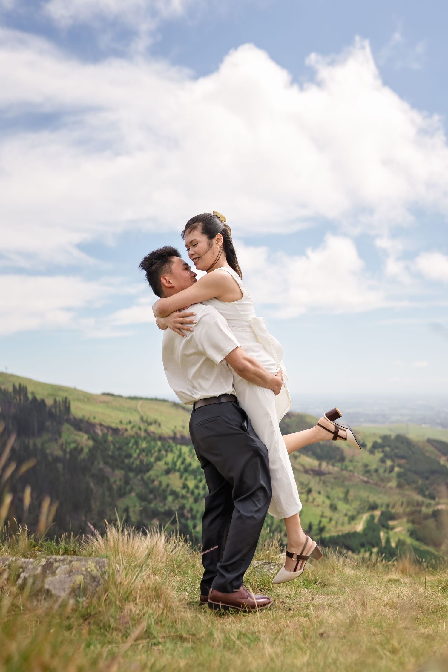 Man lifting woman in a joyful embrace on a scenic green hillside under a partly cloudy sky.