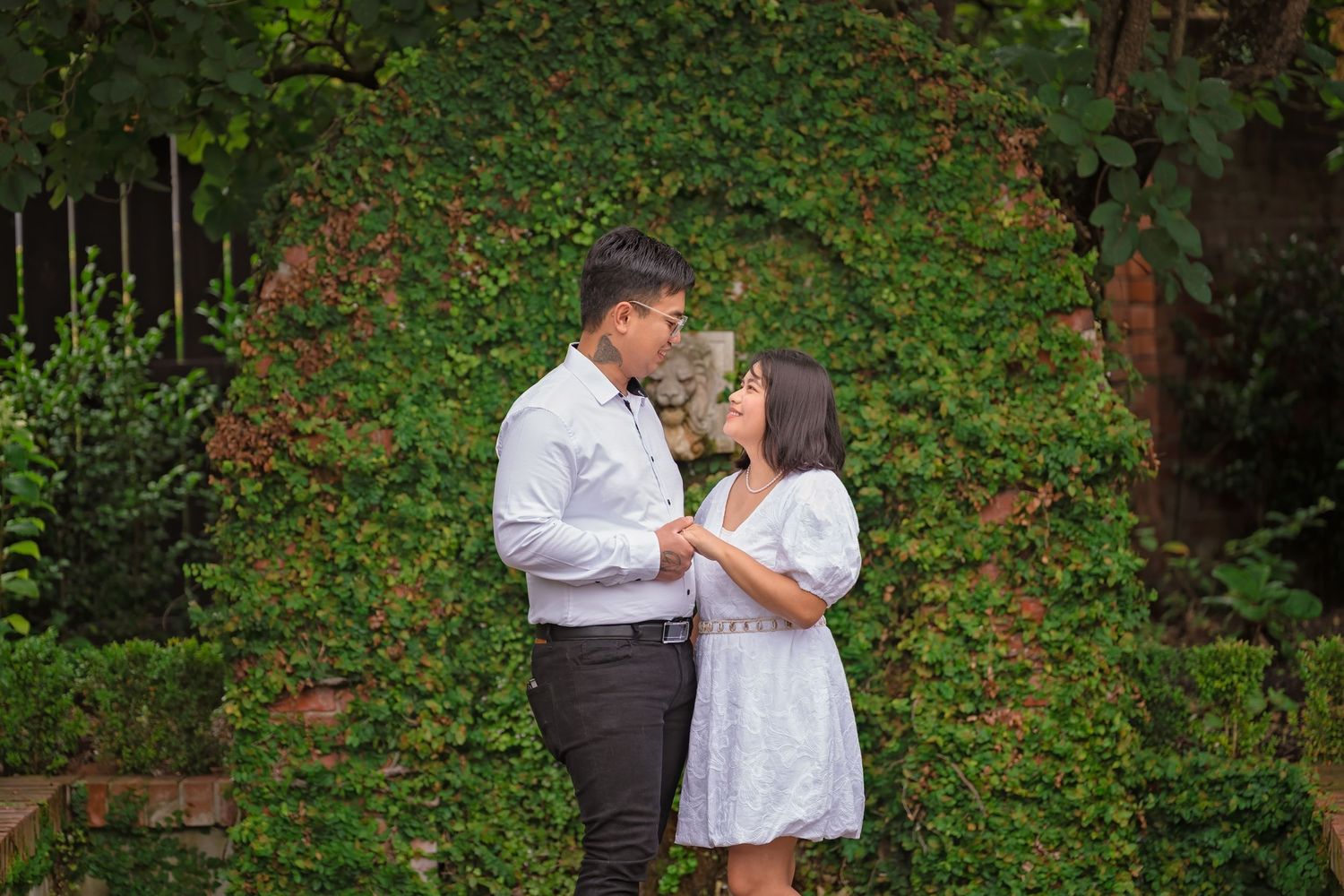 Couple holding hands in front of a lush green ivy-covered wall in a garden setting.