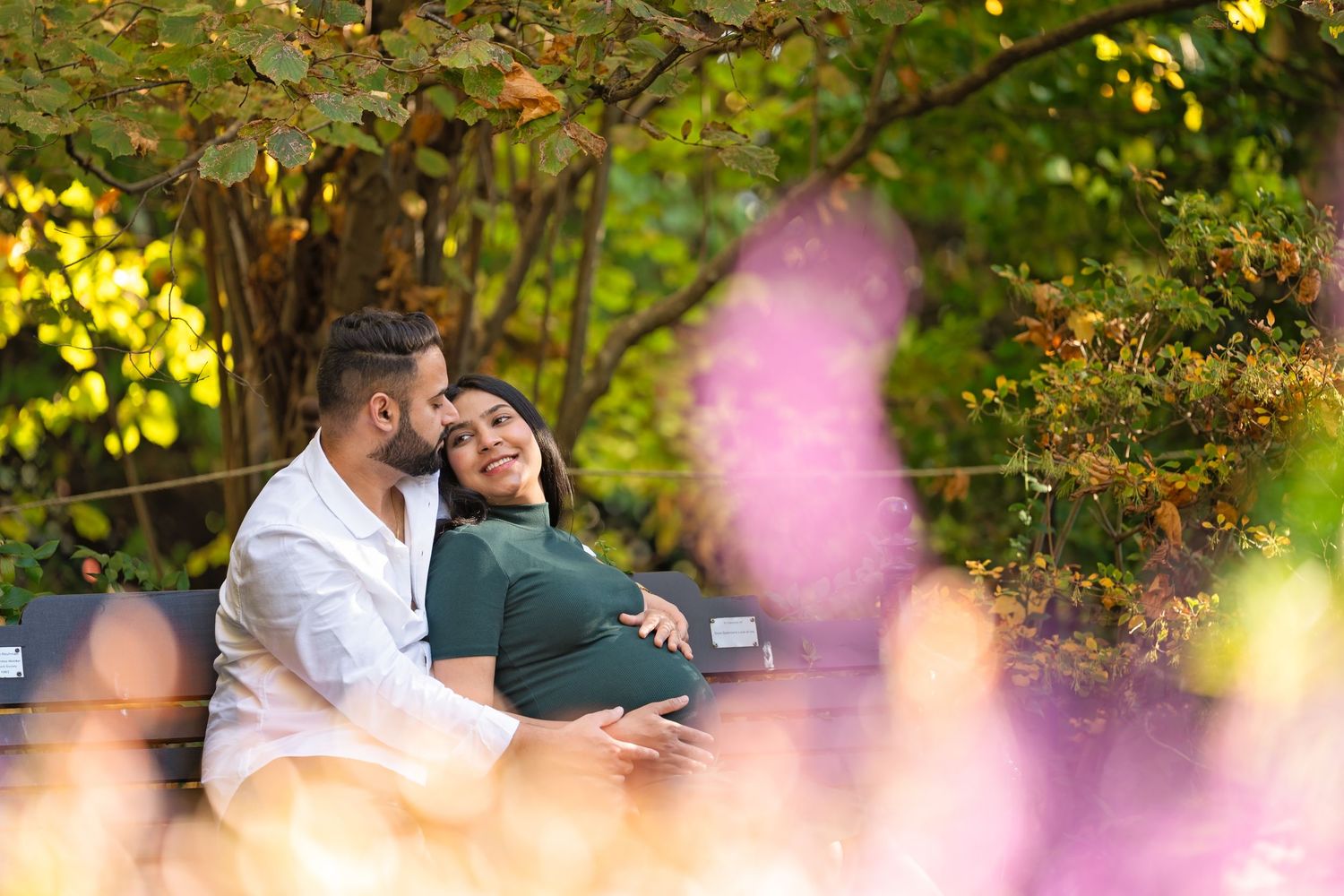 Two people sitting close together outdoors surrounded by fall foliage and soft pink bokeh lighting effects in background.