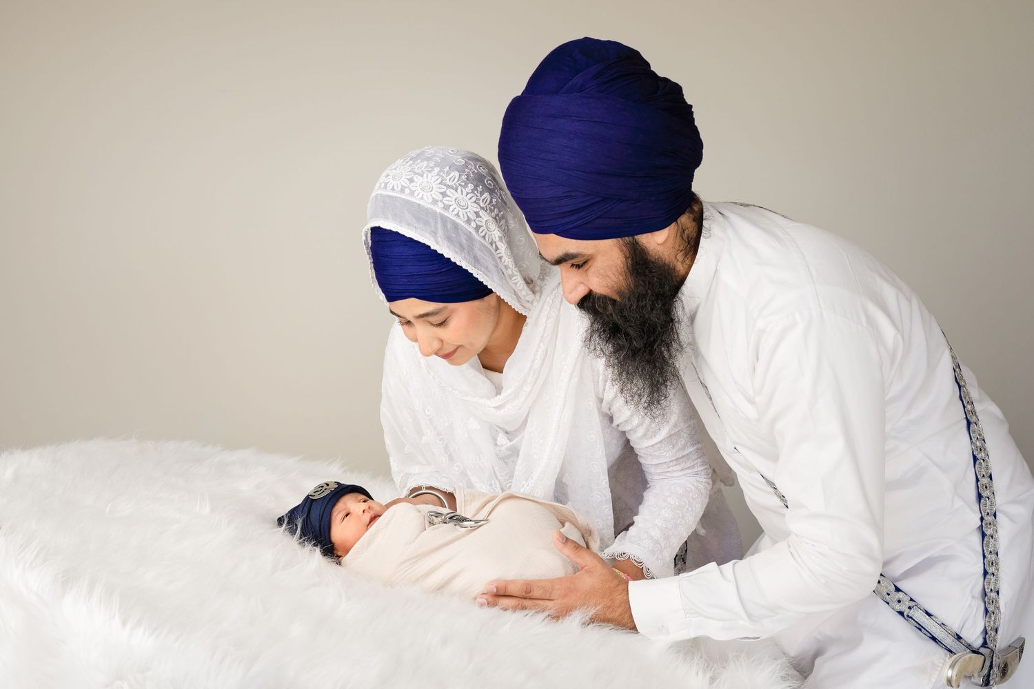 Sikh parents in traditional attire lovingly gaze at their swaddled newborn baby during a studio portrait session.