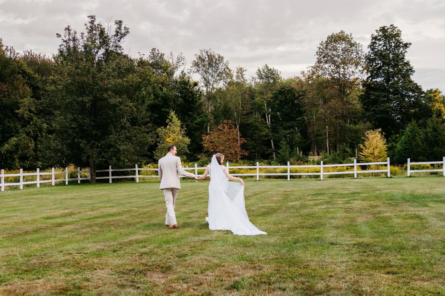A romantic wedding couple walks across a scenic green hillside with white fencing and autumn trees in the background.